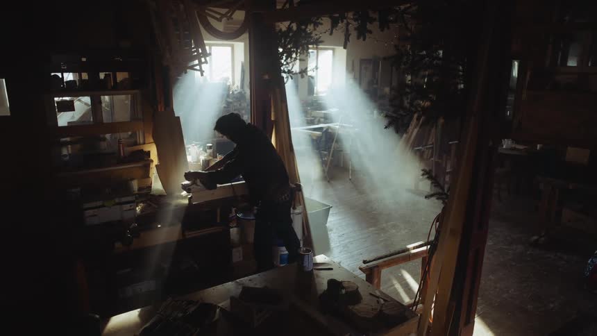 Man works in woodworking shop as sun rays brighten shadowy room