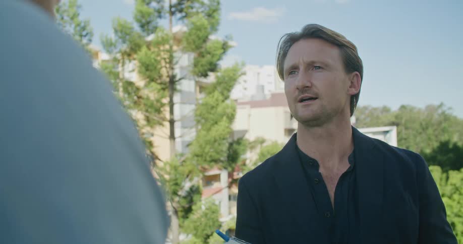 Man talks to another person while standing outside with buildings and trees in the background on a sunny day
