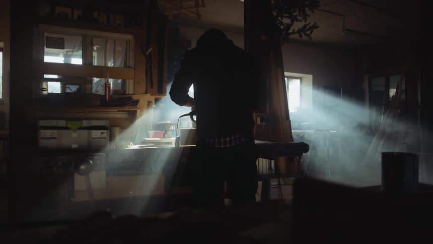 A person uses a power sander to sand a block of wood in a vice in a dimly lit workshop during the day