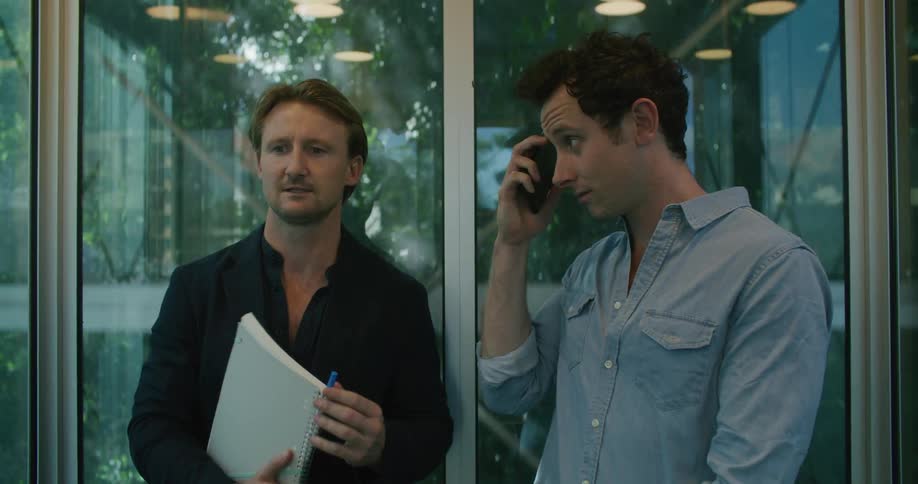 Men ride up in an elevator talking with trees and a building visible through the glass sides of the elevator