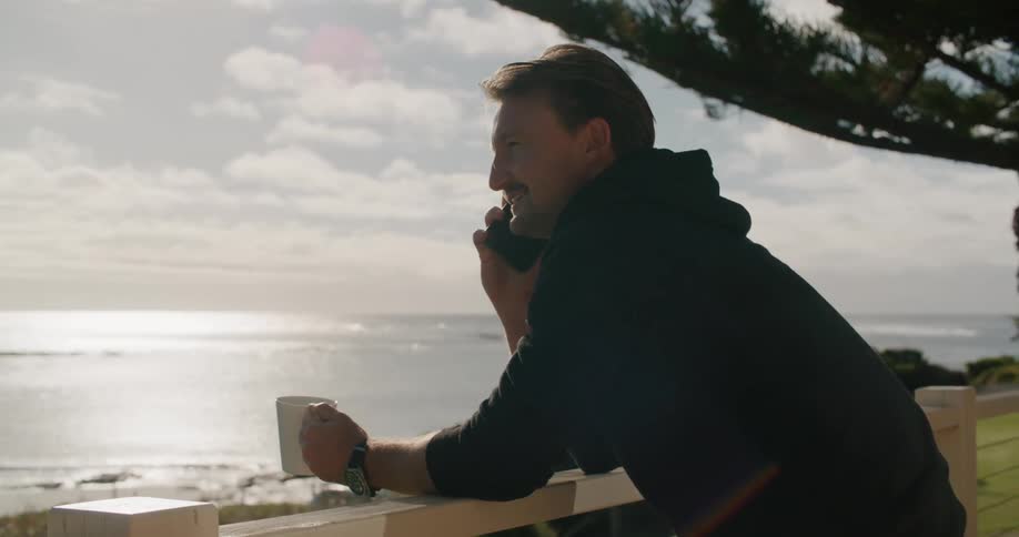 A man talks on his smartphone while leaning on a fence and looking out over the sea during the day