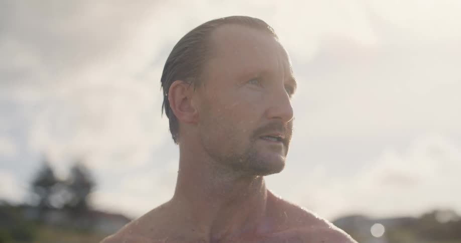 A wet shirtless man standing on a lake looking around at sunset
