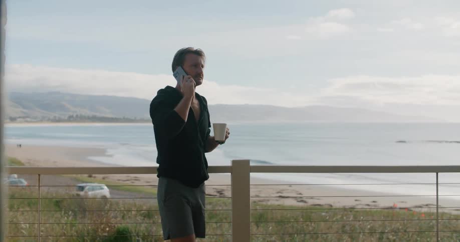 A man drinking coffee while talking on a phone near a beach