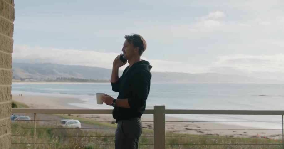 A man talks on his phone and holds a mug while walking on his balcony overlooking the beach