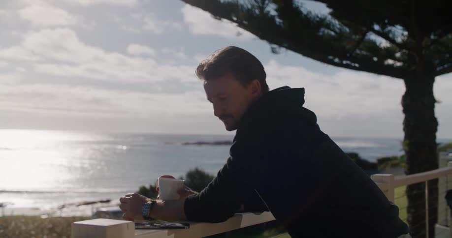 A man reclined on a railing with a cup of coffee in one hand checks his phone near the ocean