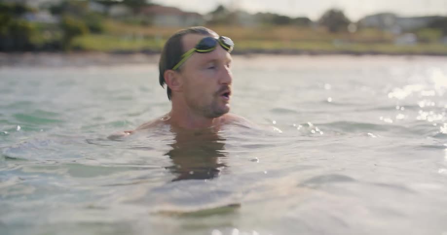 A man with goggles and a wristwatch swimming in a body of water with land the sky and clouds in the background