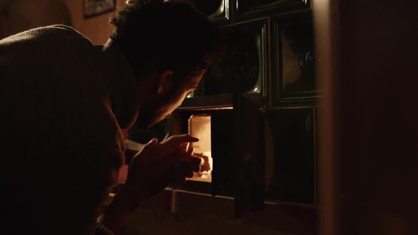 A man rubs his hands together in front of a flame in a fireplace at home