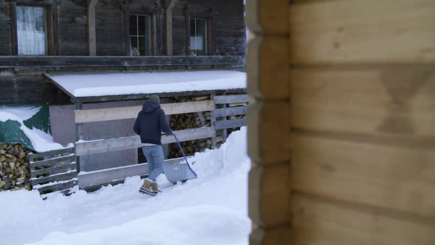 A person shoveling snow next to a wood cabin on a winter day