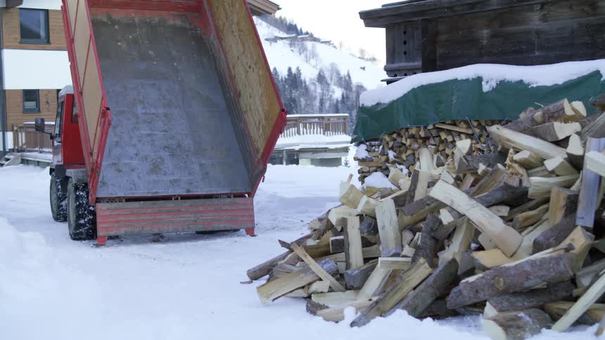 A dump truck moves away slowly from a large pile of firewood laying on the snowy ground