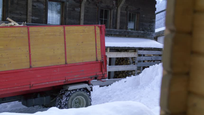 A truck loaded with split logs of firewood backs up a snow covered drive of a farmhouse