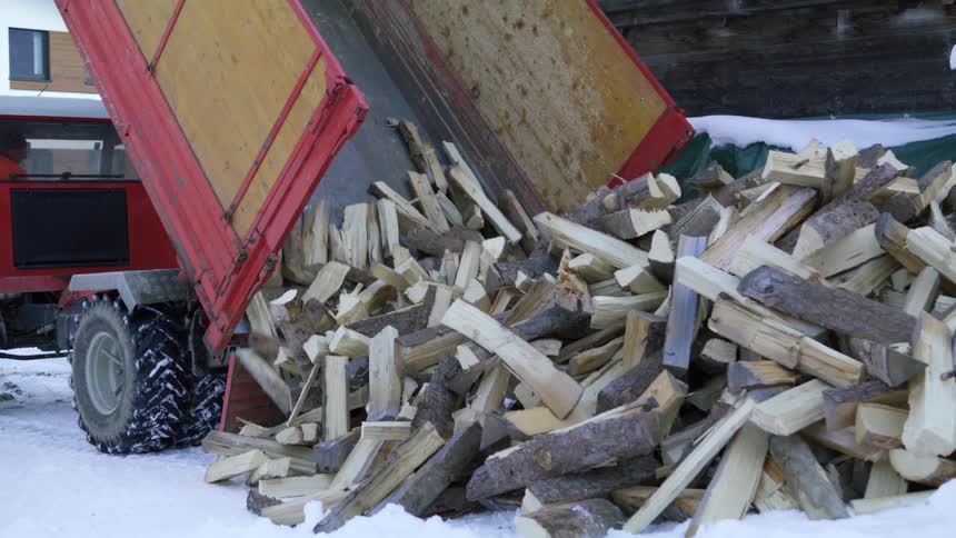 A truck dumps chopped fire wood into a pile on the snow outside a building during the day