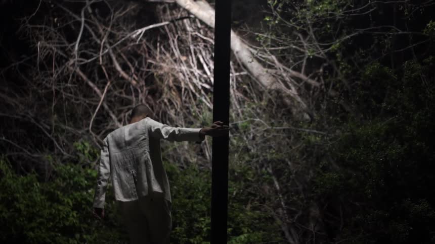 A man in a linen suits twirls around a light post at night with dense vegetation in the background