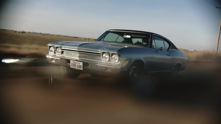 A man with sunglasses driving a car on a road next to a grassy field with the sky and clouds in the background