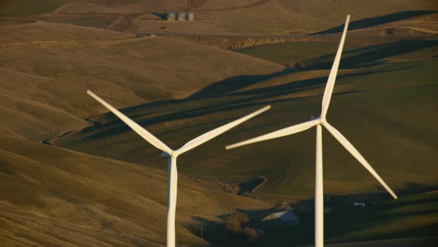Aerial shot of two wind turbines at sunset rotating pleasantly with green and tan farmland in the background
