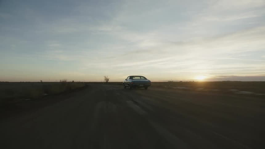 An antique car driving fast on a rural dirt road at sunset