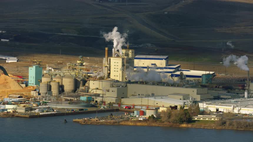 Aerial of a large factory papermill and sawmill releasing steam and exhaust emissions along the Columbia River near the Tri-Cities in Washington State