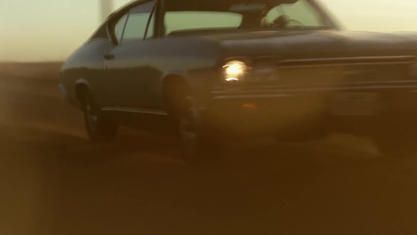 A blue vintage muscle car driving on a dusty rural road at sunset