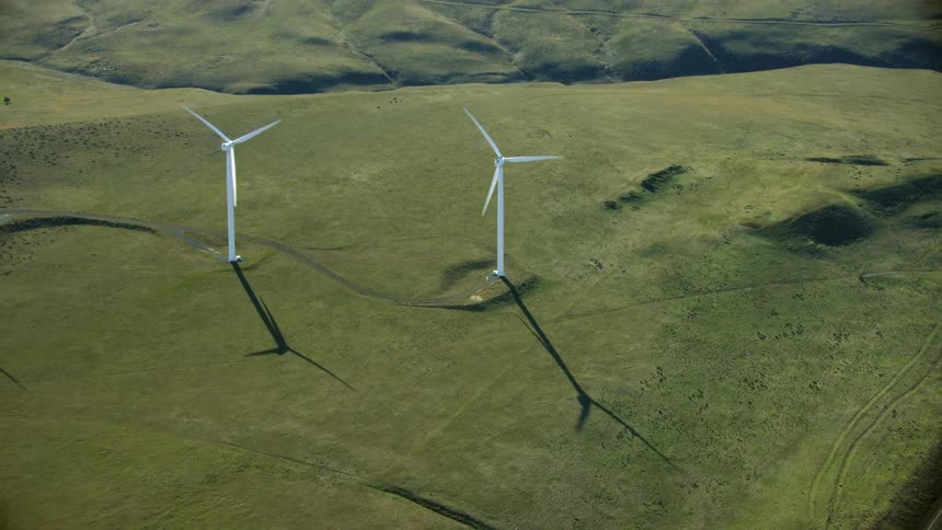 Looking down on two wind turbines as the camera tilts up to reveal a vast landscape and many more wind turbines in this aerial shot