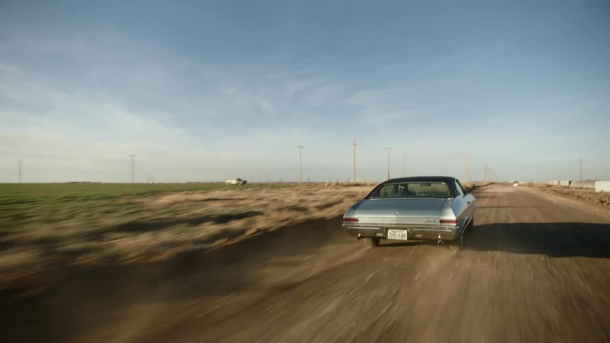 Car speeds along dirt road past green fields