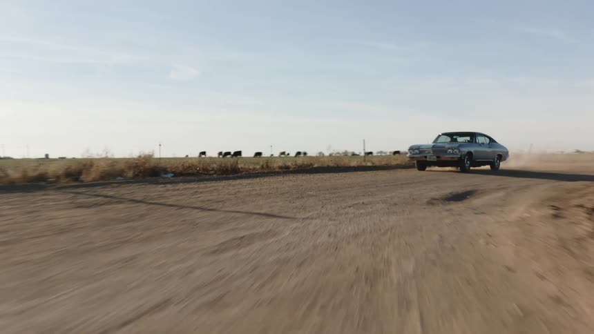 A blue vintage muscle car driving on a dusty rural road at sunset