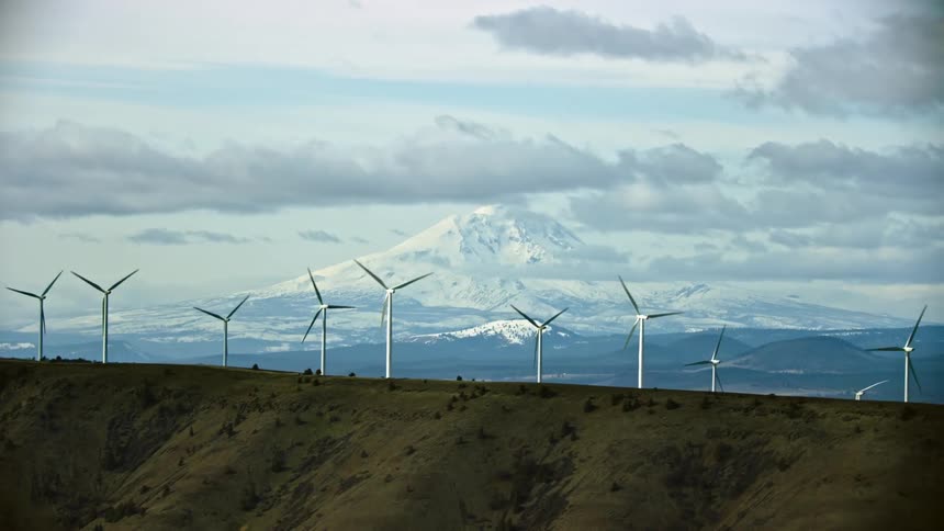 Wind farm stands on green hill with snow covered mountain on horizon under a cloudy sky with Mount Adams standing proudly in the background