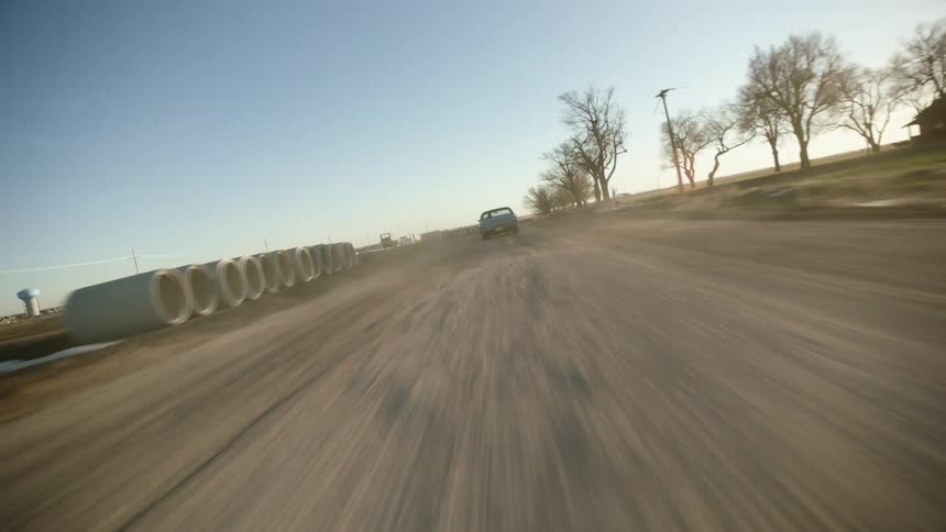 A vehicle races after another car on a dirt road past a field