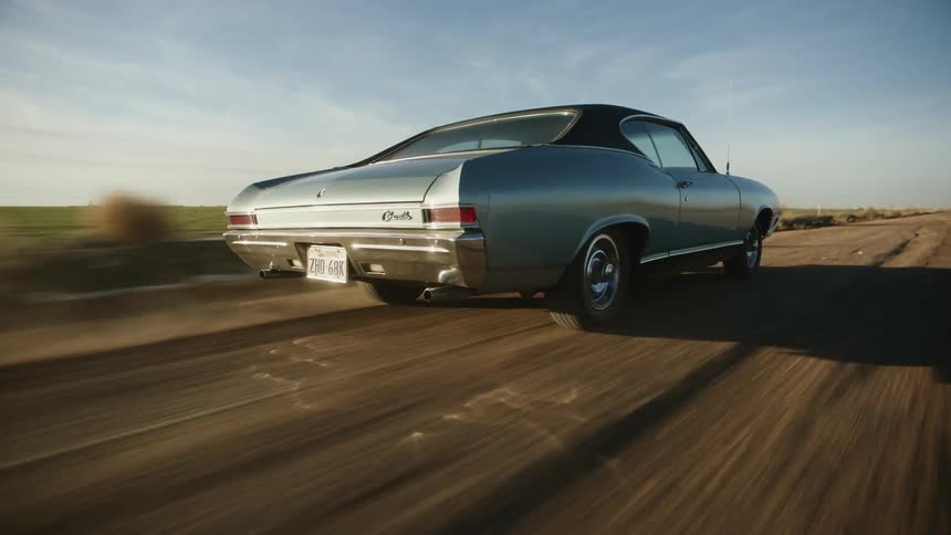 A vintage car speeds down a dirt road and past a field