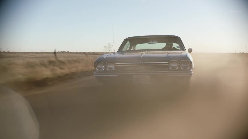 A blue vintage muscle car driving on a dusty rural road at sunset
