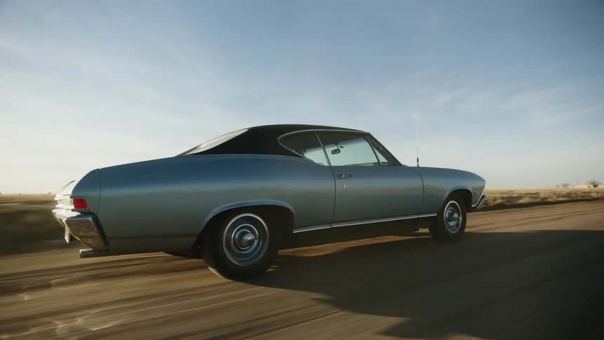 A blue vintage muscle car driving on a dusty rural road at sunset
