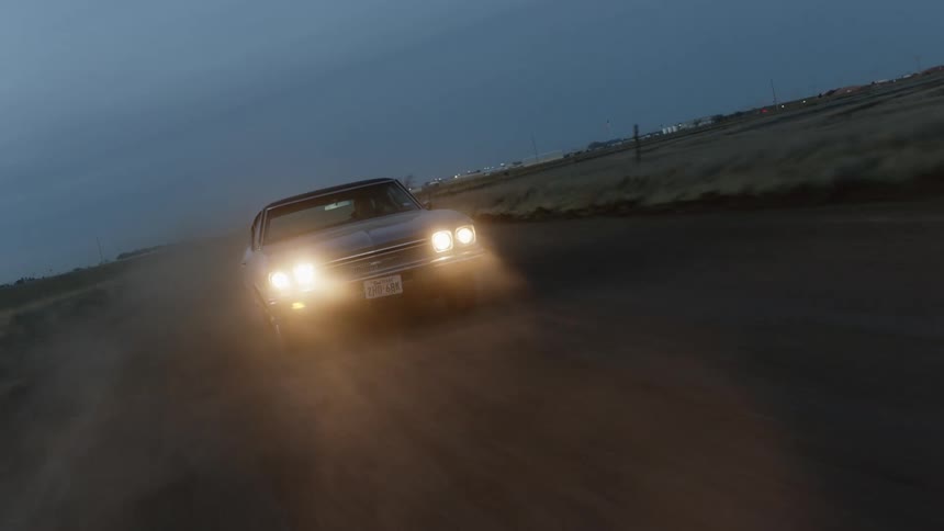 A car travels along a dirt road through the countryside on a cloudy evening
