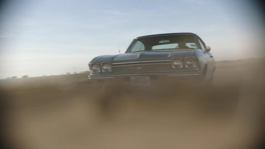 A blue vintage muscle car driving on a dusty rural road