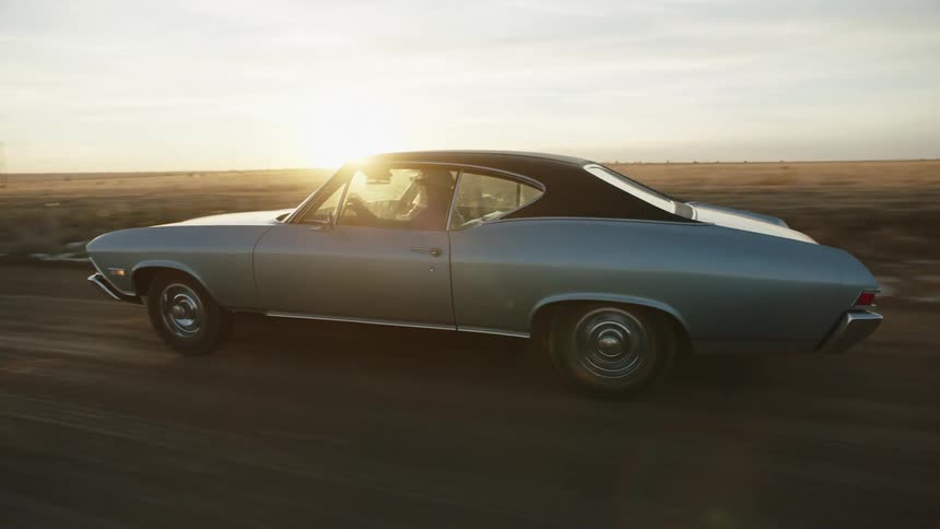 A man drives a car down a road next to a field as the sun sets