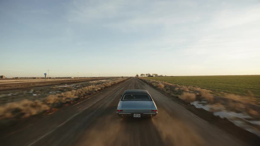 A blue vintage muscle car driving on a dusty rural road at sunset
