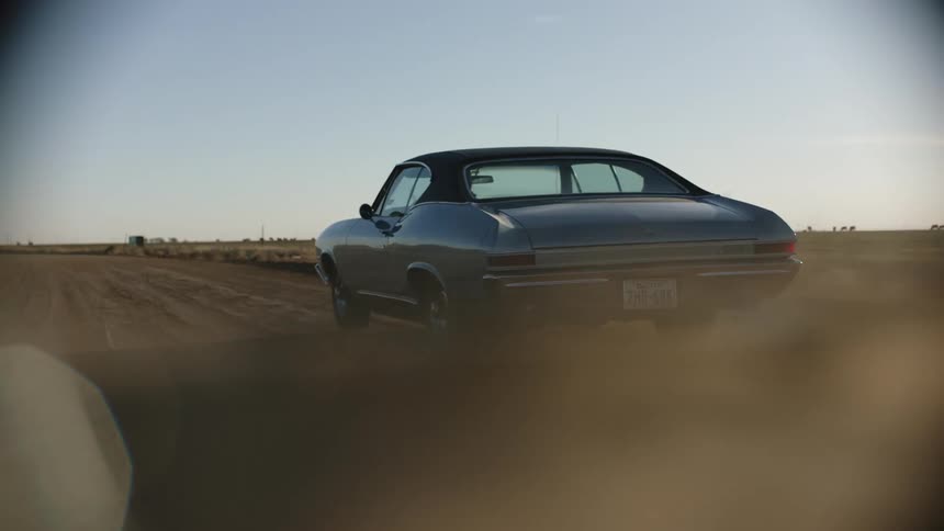 A blue vintage muscle car driving on a dusty rural road at sunset
