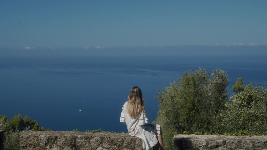 A woman is sitting on the stone wall looking at the ocean