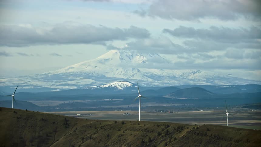Wind turbines rotate high on a ridge with a massive mountain (Mount Adams) in the background showing energy is created all over the world