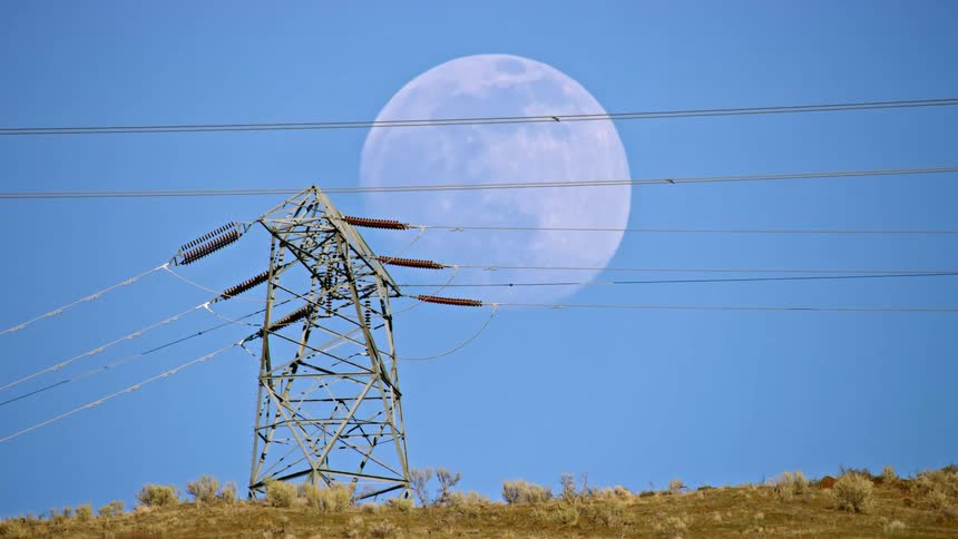 The full moon rises behind a large ridge with high tension electrical power lines and power towers in the foreground