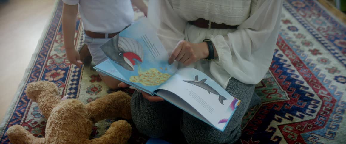 A mother sits on the floor turning pages to a book as her son sits next to her on the rug with a teddy bear by him