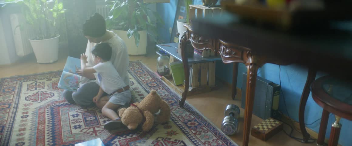 Mother and sun sit in floor on rug reading a book beside a teddy bear and a few potted plants behind them is a table and a jug to the side is a weight and a checkers board under another tall table against a blue wall
