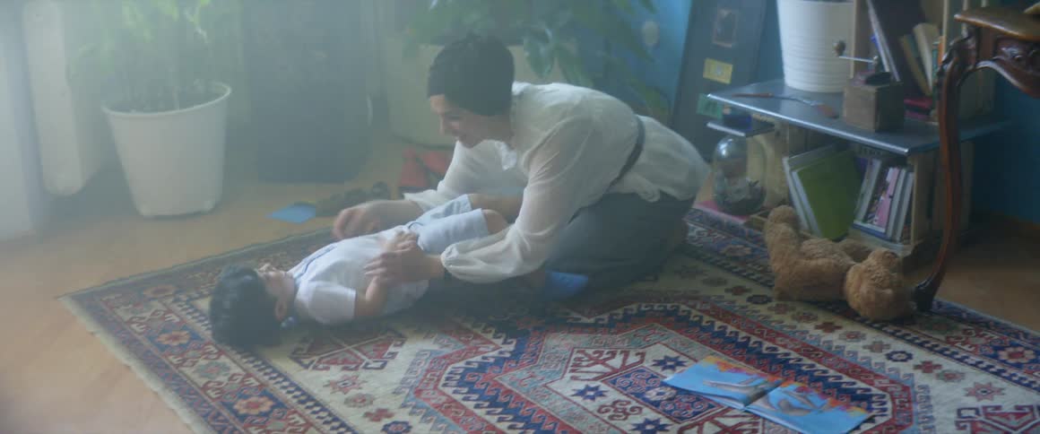 A woman playing and tickling a boy on the floor with plants in the background