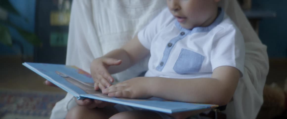 A boy sits on his moms lap as they read a book in their living room