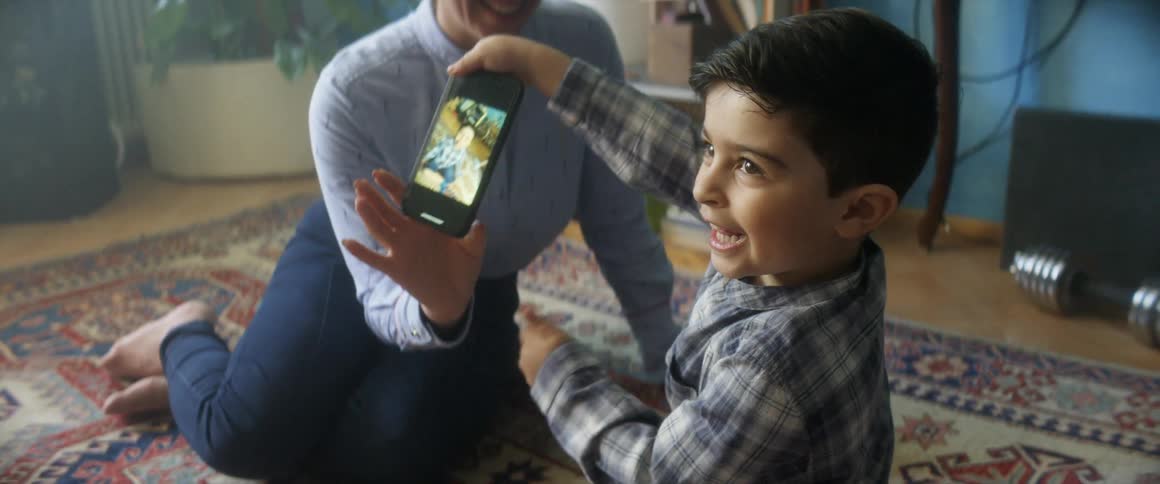 A woman shows the screen of a phone to a little boy who takes the phone and smiles as he stands up from the floor