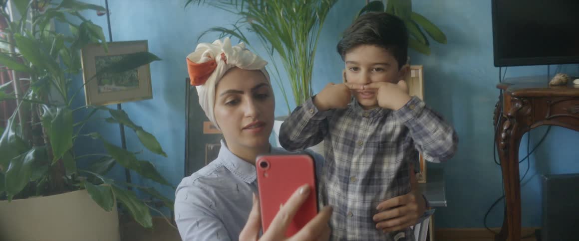 A woman holds a cell phone for a video call as her toddler boy blows kisses towards the phone