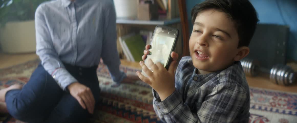 Small kid takes pictures of his mother with a smartphone sitting over a rug in a room at home