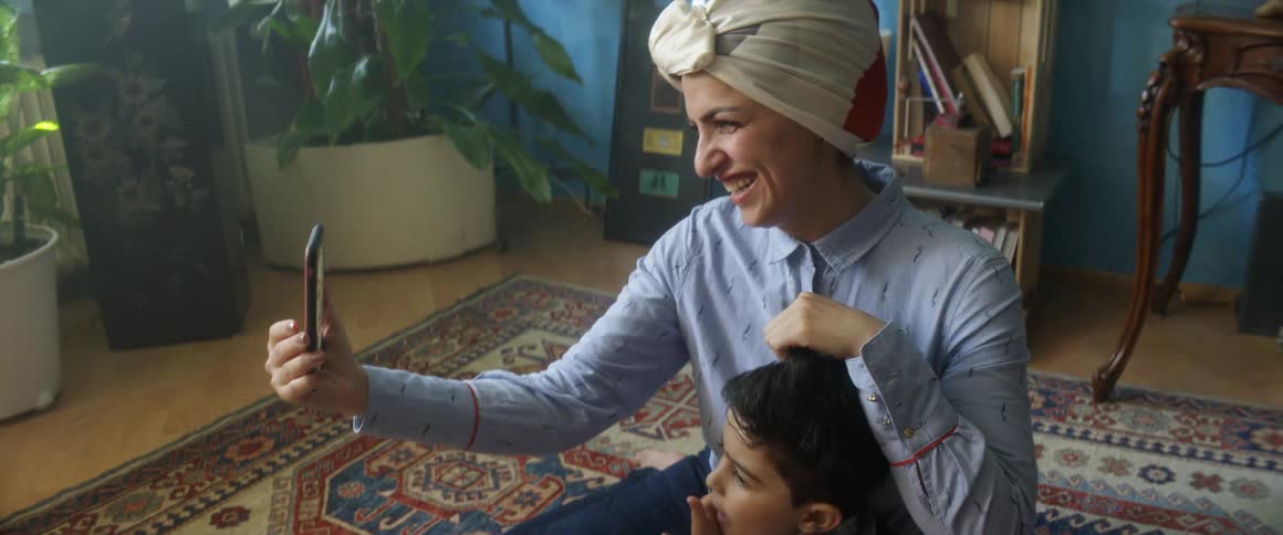 A woman at home sits on the floor with her son and they video chat on a cell phone with someone