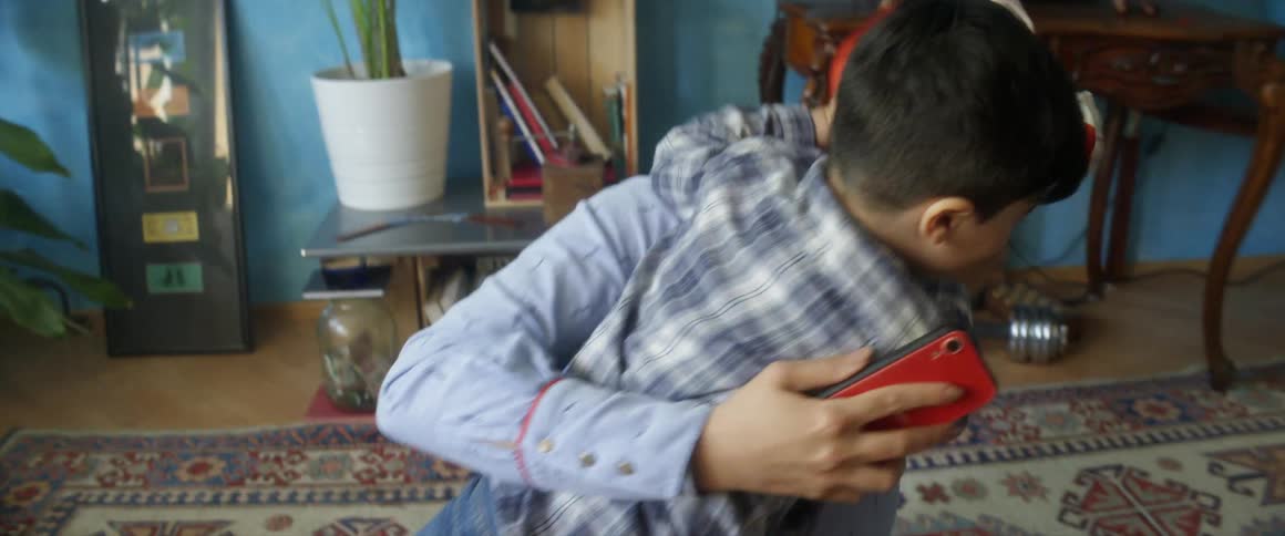 A woman with a smart phone playing with a boy in a room on the floor with plants in the background