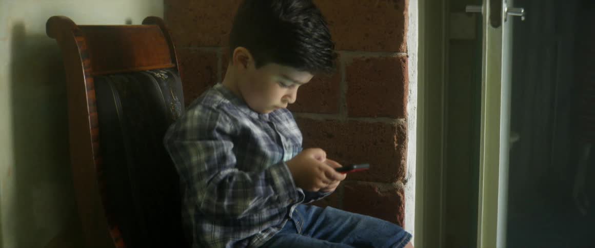 A little boy sits on a chair next to a brick wall and open door while playing on a cell phone