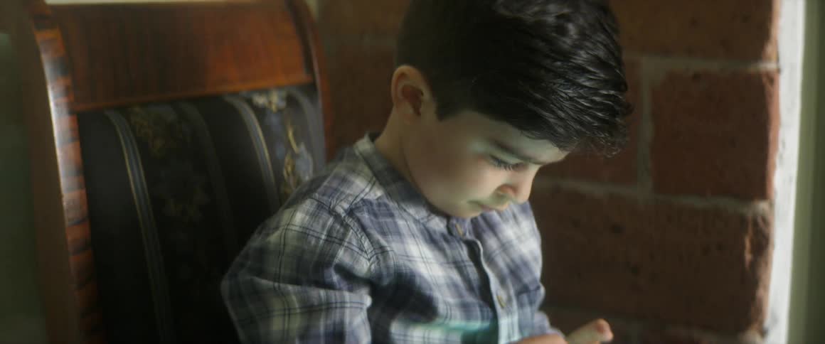 Boy sits in upholstered wooden chair near brick wall concentrating on handheld game