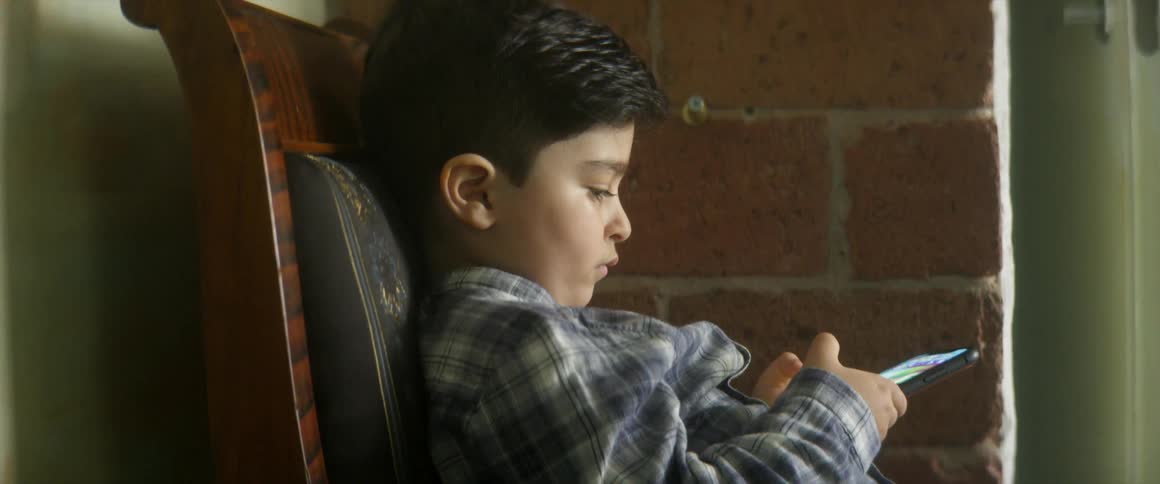 A boy sitting in a chair indoors taps the screen of a smartphone with his thumbs