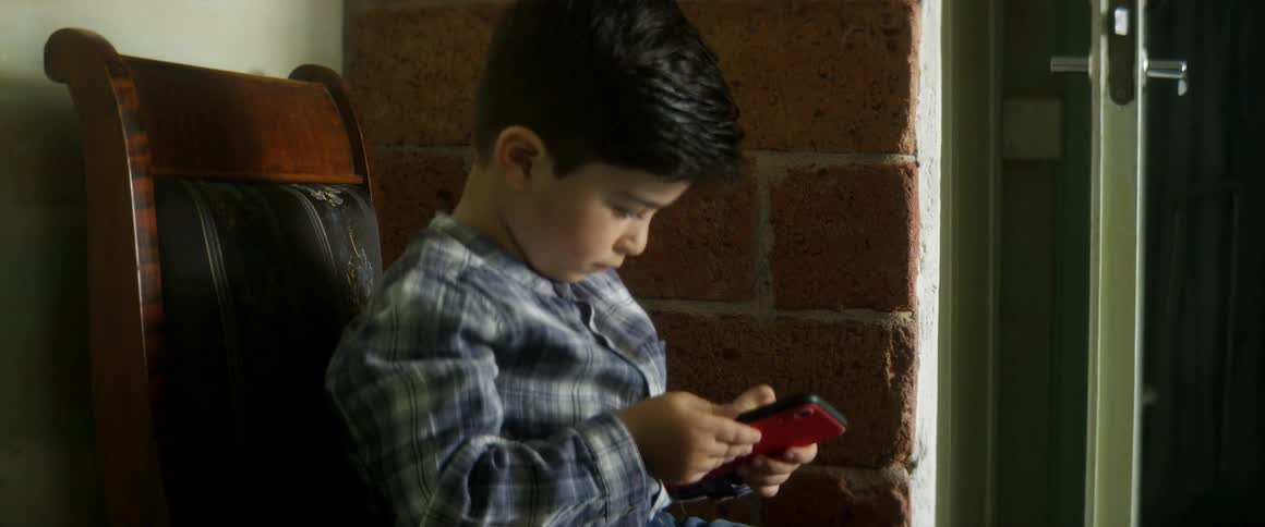 Boy sits in wooden chair beside open door and brick wall playing on smart phone